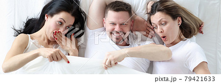 Man lying with two lovers in bed. Women looking under cover and wondering Man lying with two lovers in bed. Women looking under cover and wondering 79421633