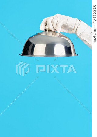 Elegant waiter's hand in white glove holding metal cloche lid cover on blue background 79445510