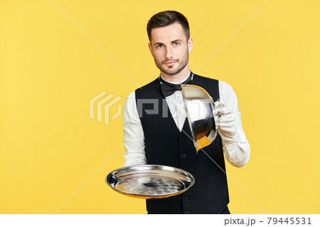 Elegant young waiter holding cloche over empty tray ready to serve on yellow background Elegant young waiter holding cloche over empty tray ready to serve on yellow background 79445531