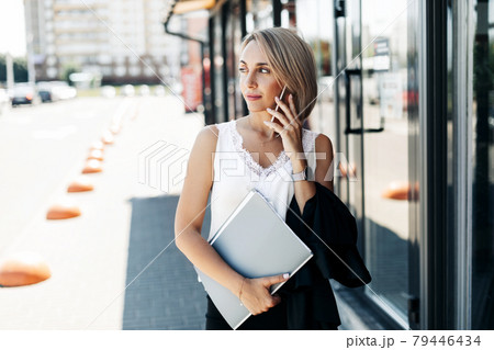 A beautiful businesswoman, talking on the phone in the city, holding a folder with documents 79446434