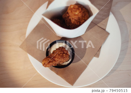 Close-up of chicken wings fried in batter. In a fast food restaurant, there is a cardboard box of chicken and sauce on the table. Sun rays fall beautifully on the hand of the girl who holds the food 79450515