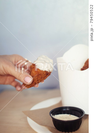 Close-up of chicken wings fried in batter. In a fast food restaurant, there is a cardboard box of chicken and sauce on the table. Sun rays fall beautifully on the hand of the girl who holds the food Close-up of chicken wings fried in batter. In a fast food restaurant, there is a cardboard box of chicken and sauce on the table. Sun rays fall beautifully on the hand of the girl who holds the food 79450522