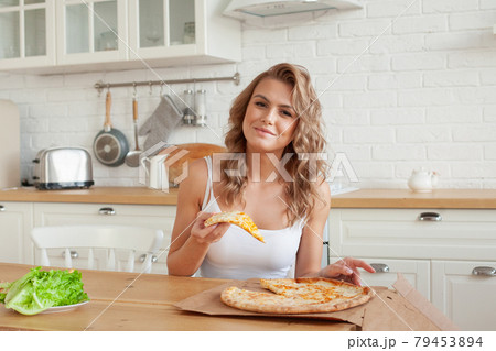 Happy blonde woman smiling and eating pizza at home Happy blonde woman smiling and eating pizza at home 79453894