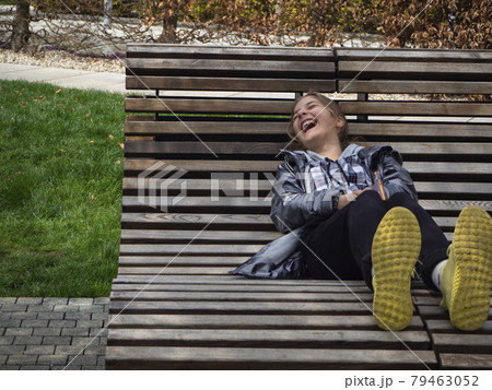 A laughing girl lies on a comfortable lounger for vacationers. A happy, joyful Girl in a jacket and yellow sneakers with a smartphone in her hands lies on a bench made of wooden planks near a green la 79463052