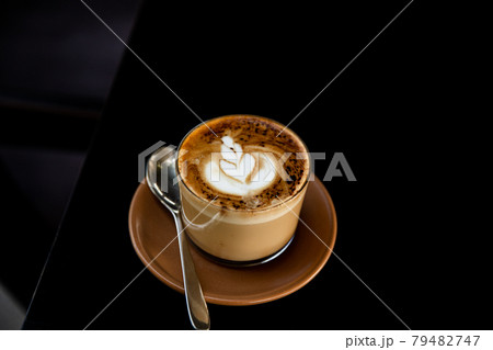 Glass of cappuccino with latte art on saucer and with spoon on the black table Glass of cappuccino with latte art on saucer and with spoon on the black table 79482747