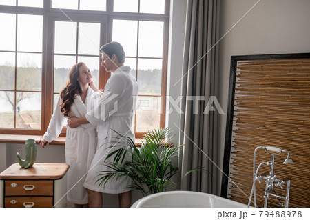 Two people in white bathrobes standing near the window and looking affectionate Two people in white bathrobes standing near the window and looking affectionate 79488978