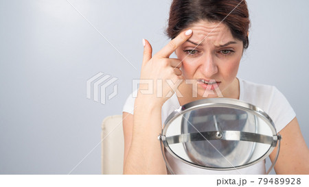 Caucasian woman looks at herself in the mirror and is upset because of facial wrinkles on her face. 79489928