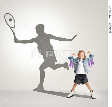 Childhood and dream about big and famous future. Conceptual image with girl and shadow of female tennis player on light gray background. Childhood and dream about big and famous future. Conceptual image with girl and shadow of female tennis player on light gray background. 79497242