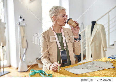 Mature seamstress with stylish short haircut drinks beverage at large cutting table in workshop 79506261