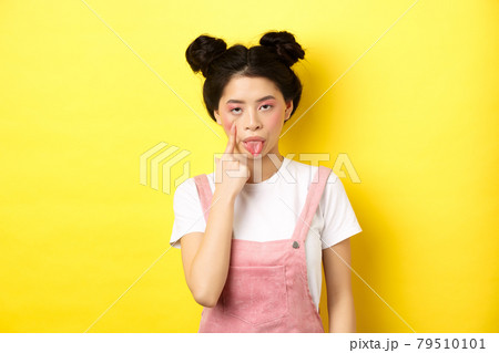 Rude asian girl stretch eyelid and showing tongue, mocking someone, standing on yellow background Rude asian girl stretch eyelid and showing tongue, mocking someone, standing on yellow background 79510101