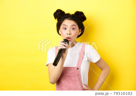 Cute asian teen girl with bright makeup, singing in microphone karaoke, standing against yellow background 79510102