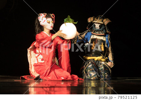 Japanese geisha in traditional kimono and fox mask holding sphere lamp and samurai warrior in armor are sitting on the knees in the dark. Traditional Japanese performance. Japanese geisha in traditional kimono and fox mask holding sphere lamp and samurai warrior in armor are sitting on the knees in the dark. Traditional Japanese performance. 79521125