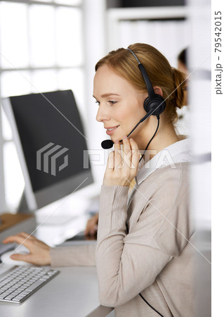 Friendly businesswoman talking by headset while sitting in office. Call center and diverse people group in business Friendly businesswoman talking by headset while sitting in office. Call center and diverse people group in business 79542015