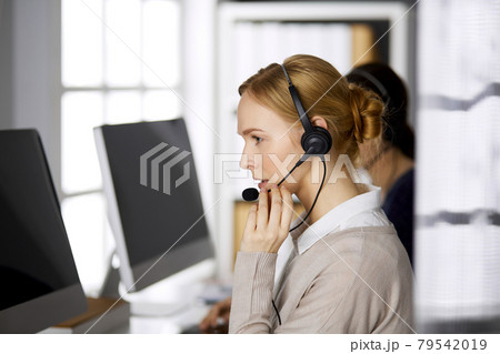 Friendly businesswoman talking by headset while sitting in office. Call center and diverse people group in business Friendly businesswoman talking by headset while sitting in office. Call center and diverse people group in business 79542019