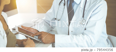 Doctor and patient discussing medical exam results while sitting at the desk in sunny clinic, close-up. Male physician using tablet computer for filling up medication history record. Doctor and patient discussing medical exam results while sitting at the desk in sunny clinic, close-up. Male physician using tablet computer for filling up medication history record. 79544356