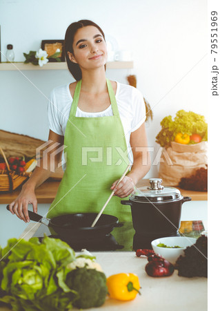 An attractive young dark-haired woman frying meal at worm pan by new keto recipe while standing and smiling in sunny kitchen. Cooking and householding concepts An attractive young dark-haired woman frying meal at worm pan by new keto recipe while standing and smiling in sunny kitchen. Cooking and householding concepts 79551969