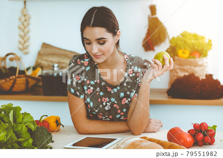 An attractive young dark-haired woman holding green apple and choosing the recipe for a delicious meal while smiling in sunny kitchen. Tablet pc is the best cookbook An attractive young dark-haired woman holding green apple and choosing the recipe for a delicious meal while smiling in sunny kitchen. Tablet pc is the best cookbook 79551982