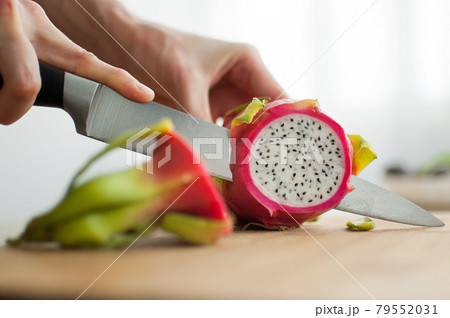 Female hands is cutting a dragon fruit or pitaya with pink skin and white pulp with black seeds on wooden cut board on the table. Exotic fruits, healthy eating concept 79552031