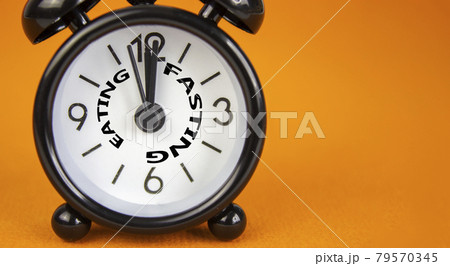 Time to fasting symbol. Beautiful black alarm clock with words 'eating fasting'. Beautiful orange background. Copy space. Healthy eating, medical and time to fasting concept. Time to fasting symbol. Beautiful black alarm clock with words 'eating fasting'. Beautiful orange background. Copy space. Healthy eating, medical and time to fasting concept. 79570345