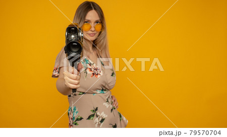 Cheerful young woman holding movie camera and posing against yellow background 79570704
