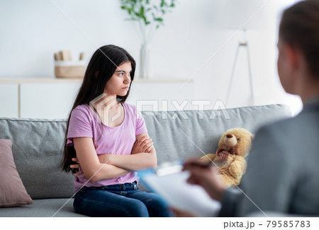 Unhappy Indian teen girl having session with psychologist at clinic, sitting with crossed arms, thinking about problems 79585783