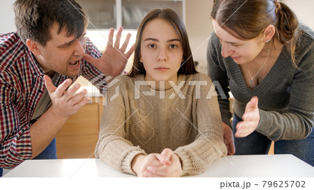 Parents yelling and shouting at teenage daughter sittin behind table and looking in camera 79625702