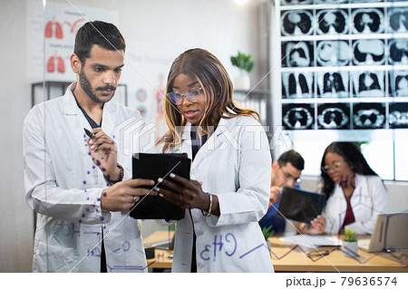 Two multiethnic male and female doctors standing behind glass wall and working on tablet pc Two multiethnic male and female doctors standing behind glass wall and working on tablet pc 79636574