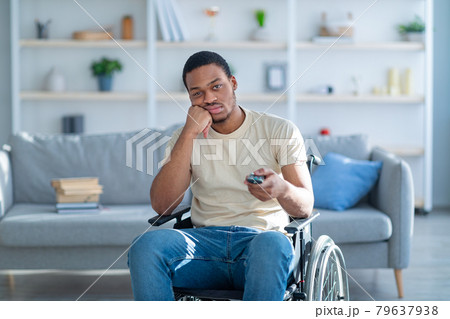 Disabled black man in wheelchair using remote control, watching dull movie on TV, cannot find interesting program Disabled black man in wheelchair using remote control, watching dull movie on TV, cannot find interesting program 79637938