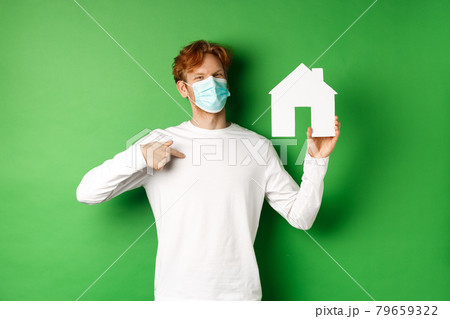 Real estate and covid-19 pandemic concept. Smiling redhead man in face mask pointing at paper house cutout, showing broker agency, standing over green background 79659322