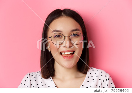 Beauty and lifestyle concept. Close-up of cute asian female model wearing trendy glasses, smiling happy at camera, standing on pink background 79659403