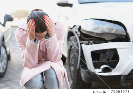 Upset young woman sits with head bowed next to wrecked car Upset young woman sits with head bowed next to wrecked car 79663573