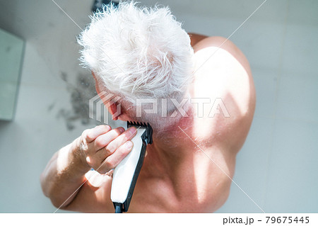 Top view white hair man doing self haircut with a clipper and looks in the mirror. Self-care at home in the conditions of global quarantine and closed hairdressers and beauty salons. electric razor Top view white hair man doing self haircut with a clipper and looks in the mirror. Self-care at home in the conditions of global quarantine and closed hairdressers and beauty salons. electric razor 79675445