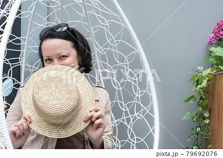 Smiling woman sits in an armchair on the summer terrace of a cafe and covers her face with a straw hat. 79692076