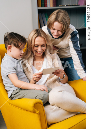 Vertical portrait of two child sons and happy mum reading surprise handmade gift card sitting in armchair Vertical portrait of two child sons and happy mum reading surprise handmade gift card sitting in armchair 79695128