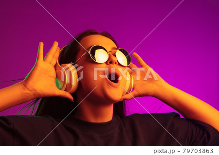 Enjoying music. Young beautiful caucasian girl in headphones and bright sunglasses isolated on pink background in neon lights 79703863