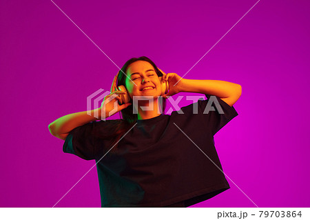 Studio image of young pretty caucasian girl with headphones listening to music isolated over pink background in neon lights 79703864