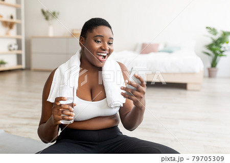 Portrait of young plus size black woman with glass of water and smartphone surfing web on break from home training Portrait of young plus size black woman with glass of water and smartphone surfing web on break from home training 79705309