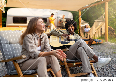Happy black guy with guitar playing music and laughing, his female friend singing song near camper van in countryside 79705363