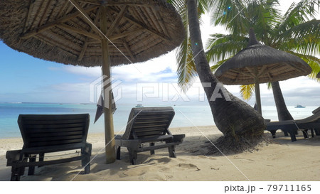 View of empty chaise-longue near native sun umbrella and palm trees against blue water, Mauritius Island View of empty chaise-longue near native sun umbrella and palm trees against blue water, Mauritius Island 79711165