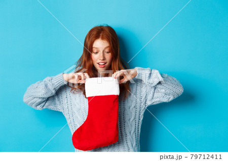 Winter holidays and gifts concept. Funny redhead girl looking inside Christmas stocking and smiling happy, receiving xmas present, standing against blue background Winter holidays and gifts concept. Funny redhead girl looking inside Christmas stocking and smiling happy, receiving xmas present, standing against blue background 79712411