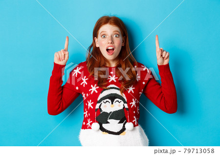 Winter holidays and celebration concept. Amazed redhead girl showing advertisement, pointing at christmas logo, standing excited over blue background Winter holidays and celebration concept. Amazed redhead girl showing advertisement, pointing at christmas logo, standing excited over blue background 79713058