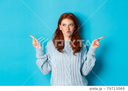 Winter holidays and people concept. Thoughtful redhead girl making decision, pointing fingers sideways, choosing between two ways, standing over blue background Winter holidays and people concept. Thoughtful redhead girl making decision, pointing fingers sideways, choosing between two ways, standing over blue background 79713060