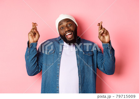 Optimistic african-american man making wish, holding fingers crossed and smiling, standing over pink background 79715009