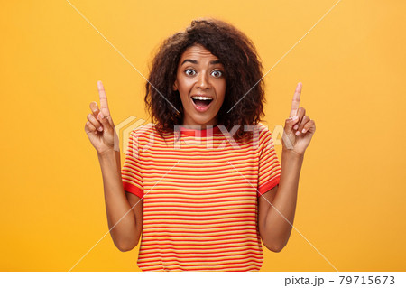 Amazed happy enthusiastic cute african american woman with curly hairstyle in striped t-shirt raising index fingers pointing up gazing mesmerized and delighted at camera over orange wall Amazed happy enthusiastic cute african american woman with curly hairstyle in striped t-shirt raising index fingers pointing up gazing mesmerized and delighted at camera over orange wall 79715673