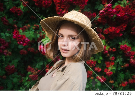 Elegant beautiful blonde young woman in a straw hat on the background of a bush of blooming roses. 79731529