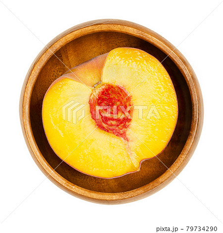 Peach half in a wooden bowl. Cross section of a ripe fruit, with yellow juicy fruit flesh, red clingstone in the center , and velvety skin. Prunus persica. Close-up from above, over white, food photo. 79734290