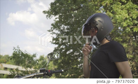 Brutal young man in sunglasses, blue sits on a custom motorcycle on the field with a black helmet. Motorcyclist on the meadow in the summer. Motorcyclist sitting near a motorcycle 79737020