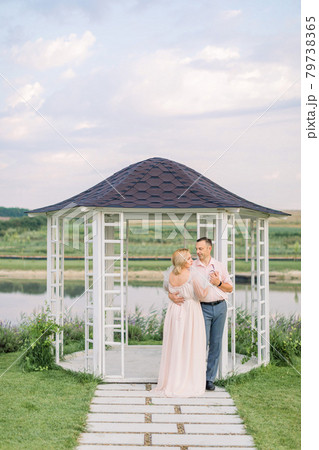 Happy mature couple in elegant clothes, standing near wooden gazebo on the shore of beautiful lake 79738365