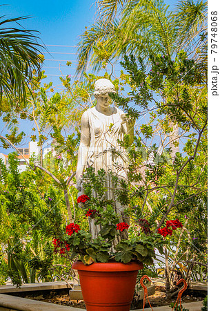 Stone statue of a woman stands in green bushes with flowers Stone statue of a woman stands in green bushes with flowers 79748068