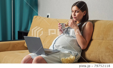 Pregnant woman eating yogurt and popcorn and looking at laptop while sitting on sofa Pregnant woman eating yogurt and popcorn and looking at laptop while sitting on sofa 79752118